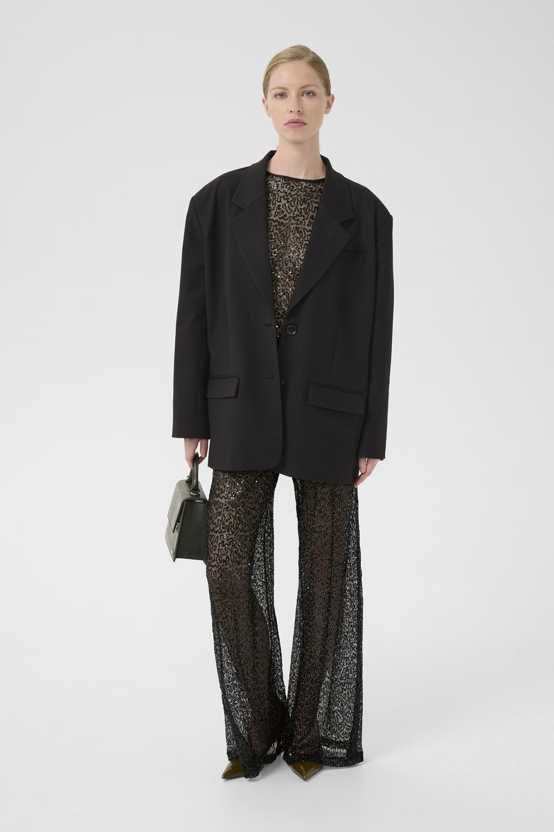 Sort oversized blazer over en palietteretop og gennemsigtige sorte blondebukser. Tilbehør inkluderer en lille grå håndtaske og spidse grønne hæle.