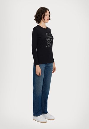 Camisa negra de manga larga con un diseño gráfico, combinada con pantalones vaqueros anchos de denim azul y zapatillas blancas. Un look simple y moderno con detalles mínimos.