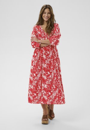 Červené šaty s kvetinovým vzorom, dlhými rukávmi, výstrihom do V a splývavou sukňou. Majú zúžený pás a sú doplnené hnedými sandálmi na platforme.