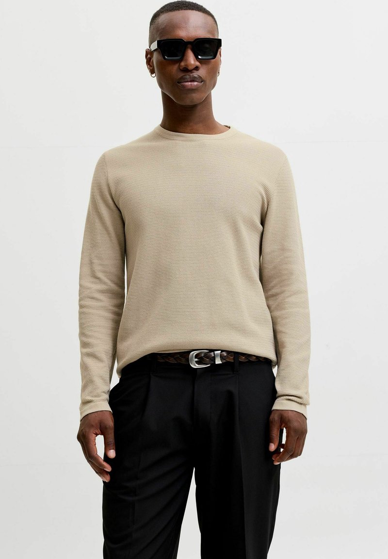 Homme portant une chemise beige à manches longues, pantalon noir, ceinture tressée, lunettes de soleil noires et petites boucles d’oreilles en créoles, debout devant un fond uni.