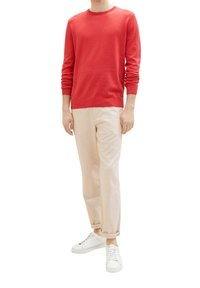 Homme portant un pull rouge à manches longues, un pantalon beige avec des revers, et des baskets blanches, debout devant un fond blanc.