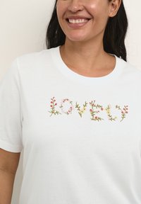 Smilende kvinde iført en hvid skjorte med ordet "KÆRLIGHED" broderet i farverige blomsterdesigns over brystet.