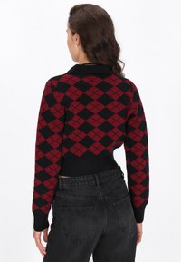 Sveter s červeným a čiernym argyle vzorom, doplnený golierom a rebrovaným lemom, v kombinácii s čiernymi nohavicami s vysokým pásom. Matná textúra, tesný strih.