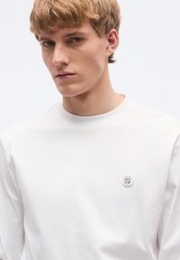 Fehér hosszú ujjú ing sima anyagból, környakú kivitelben, bal mellrészen egy kis logóval.