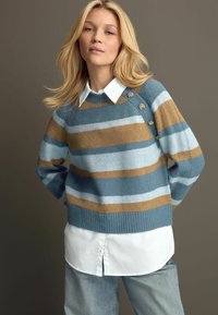 Pull rayé en mélange de laine bleu, marron et bleu clair, avec un ourlet côtelé et des accents de boutons dorés sur l'épaule, porté sur une chemise blanche à col.