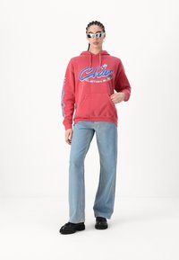 Rød hettegenser med grafisk design, med blå detaljer og kengurulomme. Kombinert med lyseblå vide jeans og svarte sko.