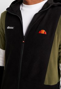 Giacca in pile nera e verde oliva con zip e cappuccio, caratterizzata da un logo ellesse e un design ricamato multicolore sul petto.