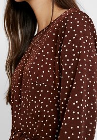 Ruskeat pilkulliset puserot, joissa on pitkät hihat, tasku, pehmeä tekstuuri ja solmittava vyötäröyksityiskohta, neutraalin taustan edessä.