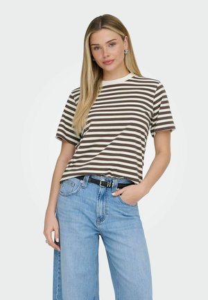 Kobieta z długimi blond włosami, ubrana w brązowo-biały prążkowany T-shirt, jasnoniebieskie dżinsy z czarnym paskiem, ręka w kieszeni, neutralne tło.