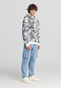Felpa bianca con stampa graffiti nera, tasca a marsupio e coulisse regolabile. Pantaloni cargo azzurri chiari con più tasche. Sneakers bianche.