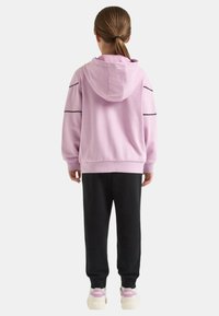 Felpa con cappuccio rosa chiaro con dettagli neri, abbinata a pantaloni della tuta neri e sneakers bianche. Il tessuto sembra morbido e casual.