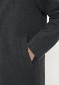 Cappotto in lana grigio scuro con una texture morbida, dotato di una tasca laterale e di una vestibilità comoda. Le estremità delle maniche sono leggermente larghe per il comfort.