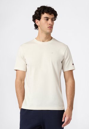 Junger Mann mit lockigem Haar, der ein schlichtes, elfenbeinfarbenes Kurzarmshirt und dunkle Hosen trägt. Er schaut nach links vor einem einfarbigen Hintergrund.