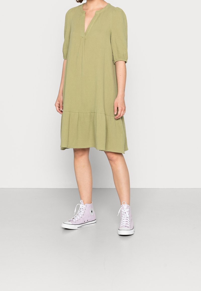 Personne portant une robe vert olive mi-longue aux genoux avec des manches trois-quarts et un ourlet à volants, associée à des baskets montantes violet clair.