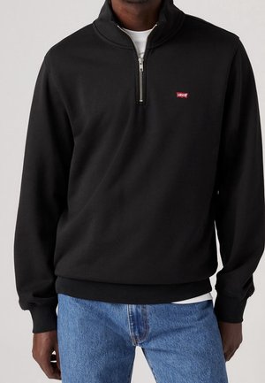Homme portant un sweat-shirt noir Levi’s à demi-fermeture éclair avec un patch logo rouge, un sous-vêtement blanc et un jean bleu, debout devant un fond uni.