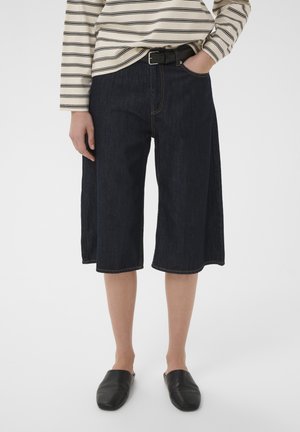 Personne portant un short large en jean foncé jusqu'aux genoux, une chemise rayée manches longues rentrée, une ceinture noire et des chaussures noires sans lacets.