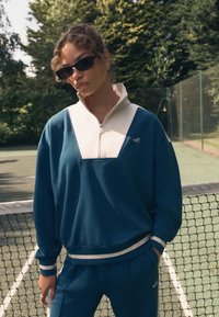 Felpa verde teal scuro con mezza cerniera crema e design a forma di V. Presenta polsini e orlo a coste. Indossata con pantaloni abbinati. Occhiali da sole indossati.