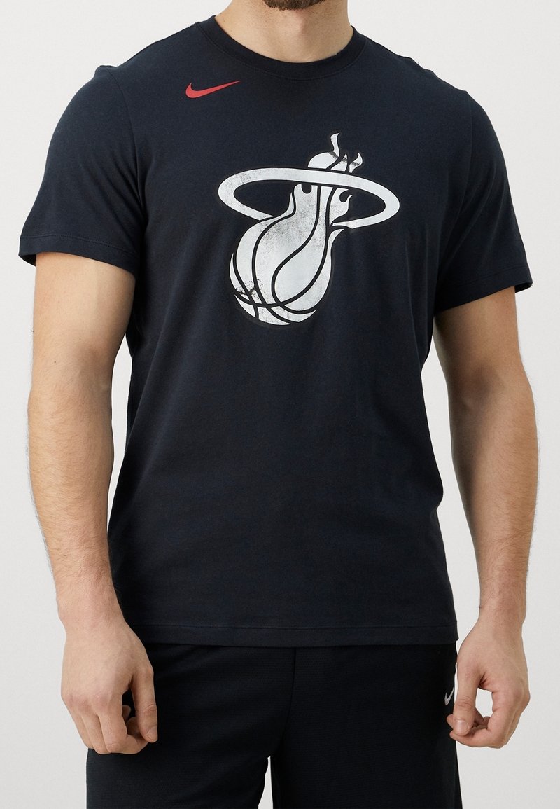 Homme portant un t-shirt noir avec le logo de l'équipe de basket Miami Heat et un petit swoosh rouge Nike sur la partie supérieure gauche de la poitrine.