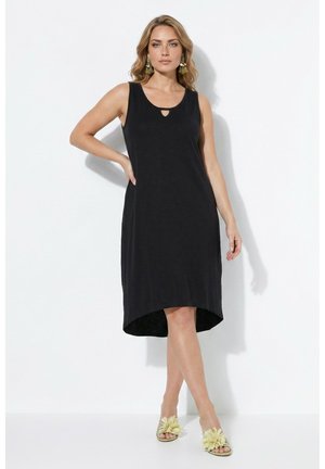 Donna in un abito nero senza maniche con orlo asimmetrico, in piedi con una mano sul fianco, indossa sandali floreali e grandi orecchini verdi.