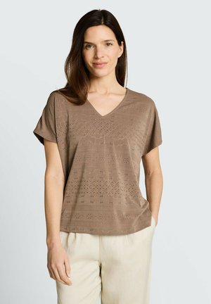 Femme aux longs cheveux bruns portant un haut marron à manches courtes à motifs avec un col en V et un pantalon beige clair, debout devant un fond uni.