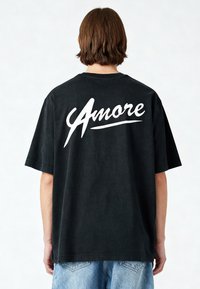 Zwarte katoenen T-shirt met korte mouwen, met het woord "Amore" in witte cursieve letters op de achterkant. Losse pasvorm en casual ontwerp.