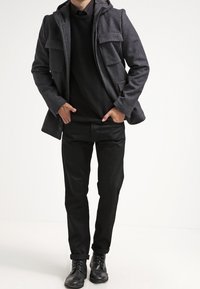 Cappotto grigio in misto lana con cappuccio, quattro tasche frontali, design a strati, indossato sopra un maglione nero e pantaloni neri, abbinato a stivali neri.