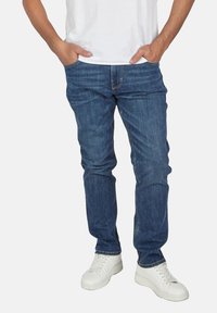 Jeans de mezclilla azul con un ajuste clásico, que presentan un estilo de cinco bolsillos y costuras sutiles, combinados con zapatillas blancas sobre un fondo en blanco.