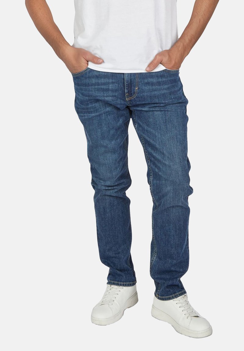 Jeans de mezclilla azul con un ajuste clásico, que presentan un estilo de cinco bolsillos y costuras sutiles, combinados con zapatillas blancas sobre un fondo en blanco.