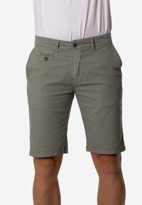 Pantalones cortos de hombre en verde oliva, hasta la rodilla, con cierre de botón y bolsillos delanteros, combinados con una camisa blanca metida dentro del pantalón.