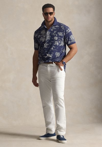 Μπλε σκουρόχρωμο polo με κοντά μανίκια και λευκό τροπικό μοτίβο, συνδυασμένο με λευκά τζιν και μπλε αθλητικά παπούτσια. Αξεσουάρ περιλαμβάνουν γυαλιά ηλίου και ρολόι.