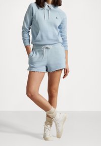 Henkilö, jolla on päällään vaaleansininen huppari ja siihen sopivat kuluneet vaaleansiniset shortsit, sekä kermahenkiset sukat ja valkoiset korkeavartiset tennarit.