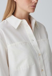 White button-up shirt featuring a classic collar, two chest pockets, and subtle ribbed texture. Button fasteners are light-colored.