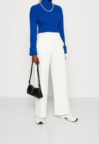 Blauwe geribbelde coltrui, witte brede broekspijpen, zwarte handtas en witte sneakers. Eenvoudig, modern ontwerp met contrasterende kleuren.