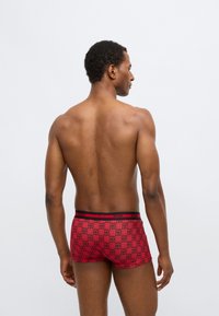 Κόκκινα κοντά ανδρικά εσώρουχα με μοτίβο πλέγματος και λογότυπα "HUGO". Η ζώνη μέσης είναι μαύρη με κόκκινα γράμματα. Κατασκευασμένα από απαλό, ελαστικό ύφασμα.