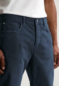 Pantalones de pana azul marino con un acabado texturizado, que cuentan con una cremallera, bolsillos frontales y un diseño ajustado.