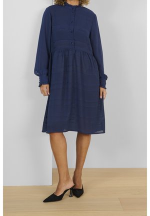 Frau trägt ein marineblaues Midikleid mit langen Ärmeln und Knöpfen sowie schwarze spitze Mule mit Absatz, steht auf hellem Holzboden.