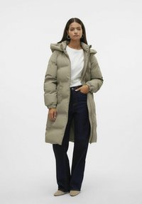 Manteau matelassé vert olive long avec capuche, doté d'une fermeture éclair et de poignets élastiques, associé à un jean large bleu foncé et des chaussures beiges.