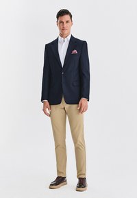 Granatowa marynarka z dwoma przednimi kieszeniami, biała koszula, beżowe spodnie oraz ciemne buty. Różowa poszetka dodaje kontrastującego akcentu.