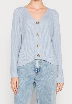 Cardigan en maille côtelée bleu clair avec quatre boutons en bois, col en V, manches longues, porté avec un jean taille haute délavé clair.