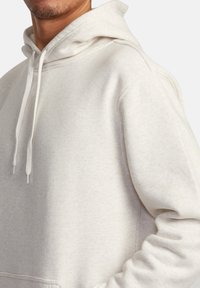 Hoodie beige clair avec une texture douce, des cordons de serrage et une poche kangourou. Il présente une coupe décontractée et des détails cousus sur les coutures.