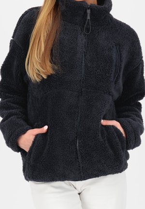 Femme portant une veste en polaire noire zippée avec les mains dans les poches avant, assortie à un pantalon de couleur claire.