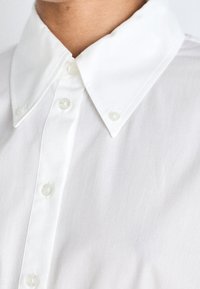 Camicia bianca con colletto dalla texture liscia, dotata di bottoni trasparenti e design a bottoni, che enfatizza uno stile minimalista e la semplicità.