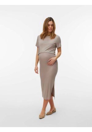 Rase naine beežis soonikkoes lühikeste varrukatega tipus ja sobiva midi pikkusega seelikuga, seisab ja hoiab õrnalt kõhtu valgel taustal.