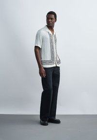 Hombre de pie con una camisa blanca de manga corta con estampado, pantalones negros y zapatos negros, mirando de lado sobre un fondo liso.