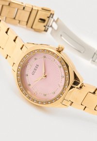 Reloj de metal dorado con esfera rosa en efecto rayos de sol, detalles de cristal y manecillas doradas. Cuenta con una correa de brazalete metálica dorada.