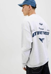 Sudadera gris claro de corte suelto, con puños de canalé y texto impreso en azul. Combinada con jeans oscuros. Gorra azul llevada hacia atrás.