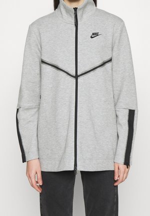 Osoba nosząca jasnoszarą kurtkę Nike na zamek z czarnymi paskami na rękawach i klatce piersiowej, w połączeniu z ciemnymi spodniami.
