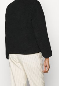 Czarna, strukturalna polarowa kurtka z długimi rękawami. Prosty, prosty fason; noszona na kremowe, w prążki spodnie. Bez widocznych elementów metalowych ani akcentów.