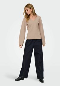 Femme portant un pull beige à col en V, un pantalon large noir et des bottines noires, debout en regardant vers le bas à sa gauche.