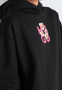 Sudadera negra con un llamativo logo "HUGO" en rosa y acentos florales. La tela parece suave, con un corte relajado y capucha con cordones.